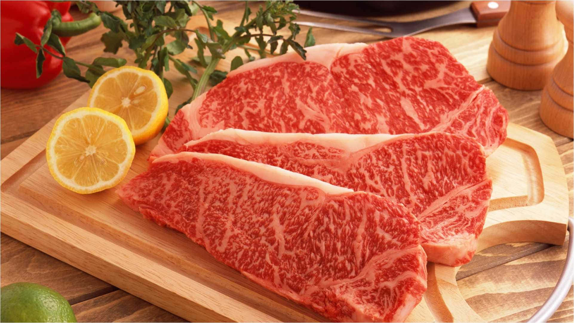 meat 桌布 壁紙 — 三塊大理石紋牛肉扒放在木砧板上，旁邊有檸檬片和新鮮香草。