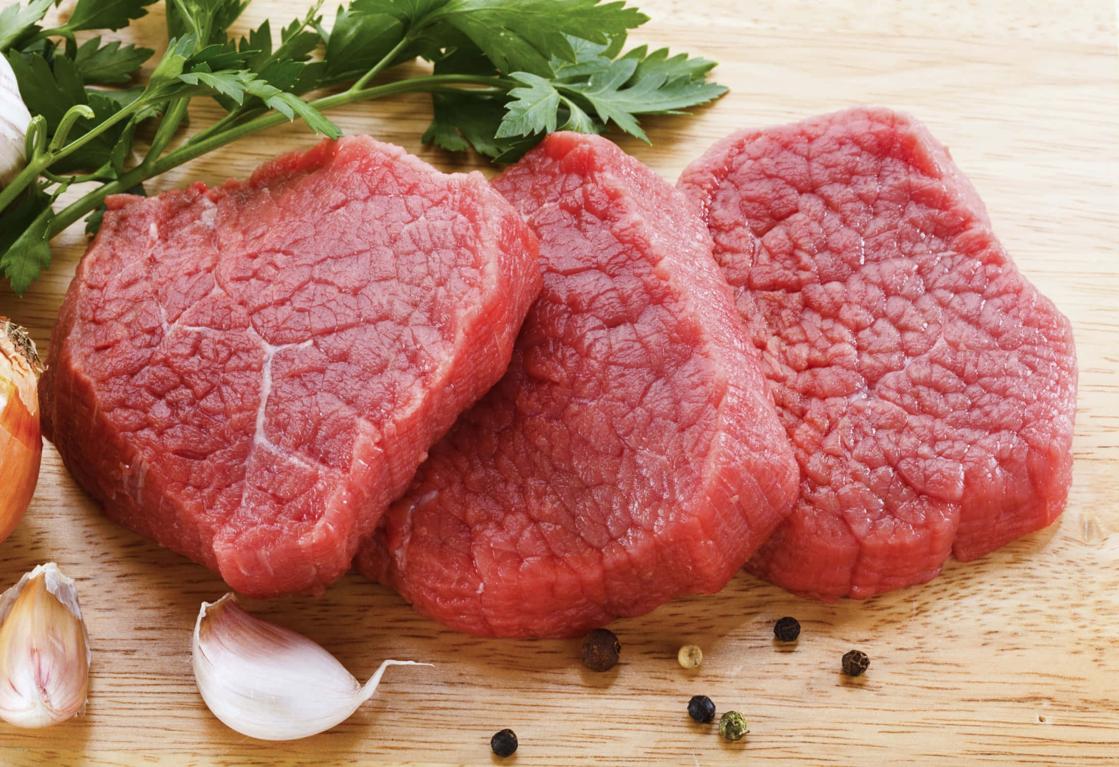 meat 桌布 壁紙 — 三塊帶有明顯大理石花紋的生牛肉扒放在木板上，旁邊是新鮮的歐芹、大蒜和胡椒粒。