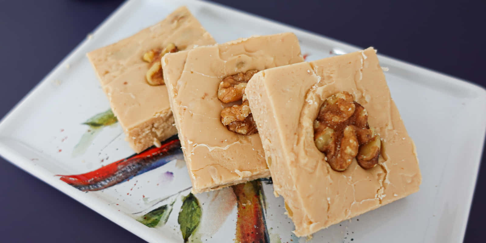 Penuche Fudge 桌布 壁紙 — 三塊方形奶油花生醬軟糖，上面放著完整的核桃，放在一個裝飾性的白色盤子上。