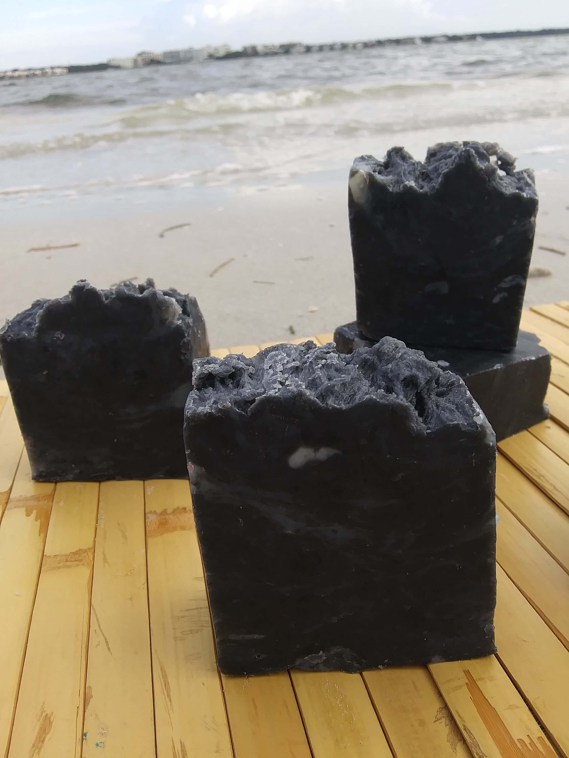 Black Soap 桌布 壁紙 — 三塊深色紋理肥皂放在木甲板上，背後有海浪拍打。