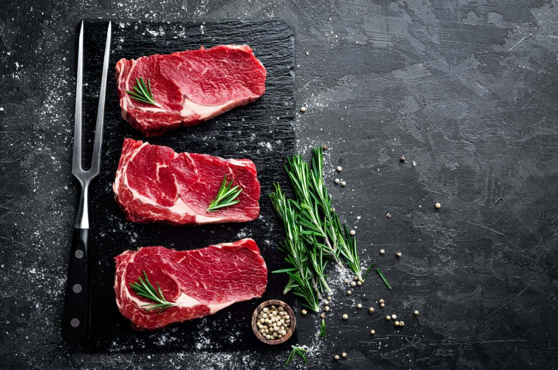Red Meat 桌布 壁紙 — 三塊生牛肉扒以迷迭香點綴，放在深色石板板上，旁邊有叉子和胡椒粒。