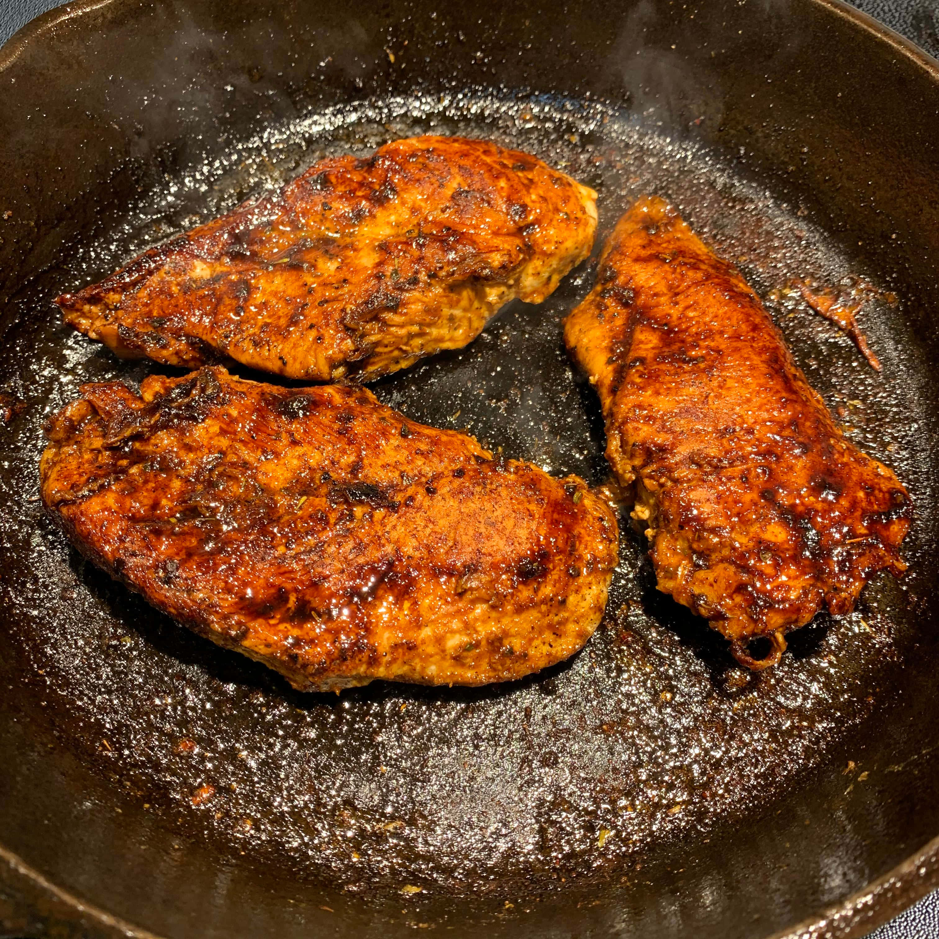 Blackened Chicken 桌布 壁紙 — 三塊調味雞胸肉在常用的鑄鐵煎鍋中滋滋作響，帶有金黃色的外皮。