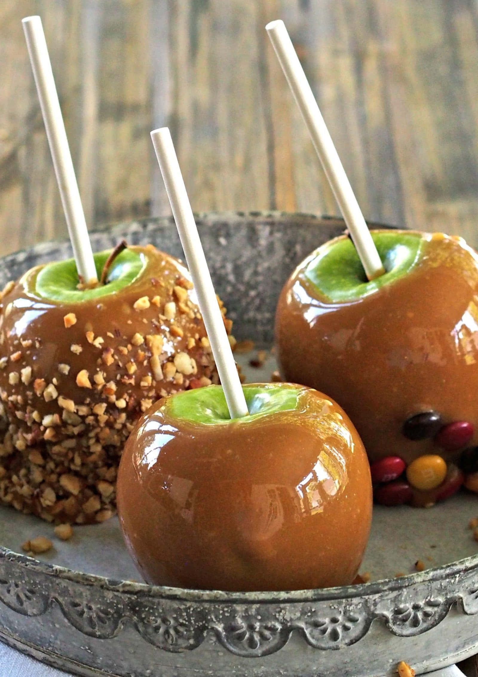 Caramel Apples 桌布 壁紙 — 三塊閃亮的焦糖蘋果，附有棍子和碎堅果，放在復古金屬托盤上，背景是質樸的木質背景。
