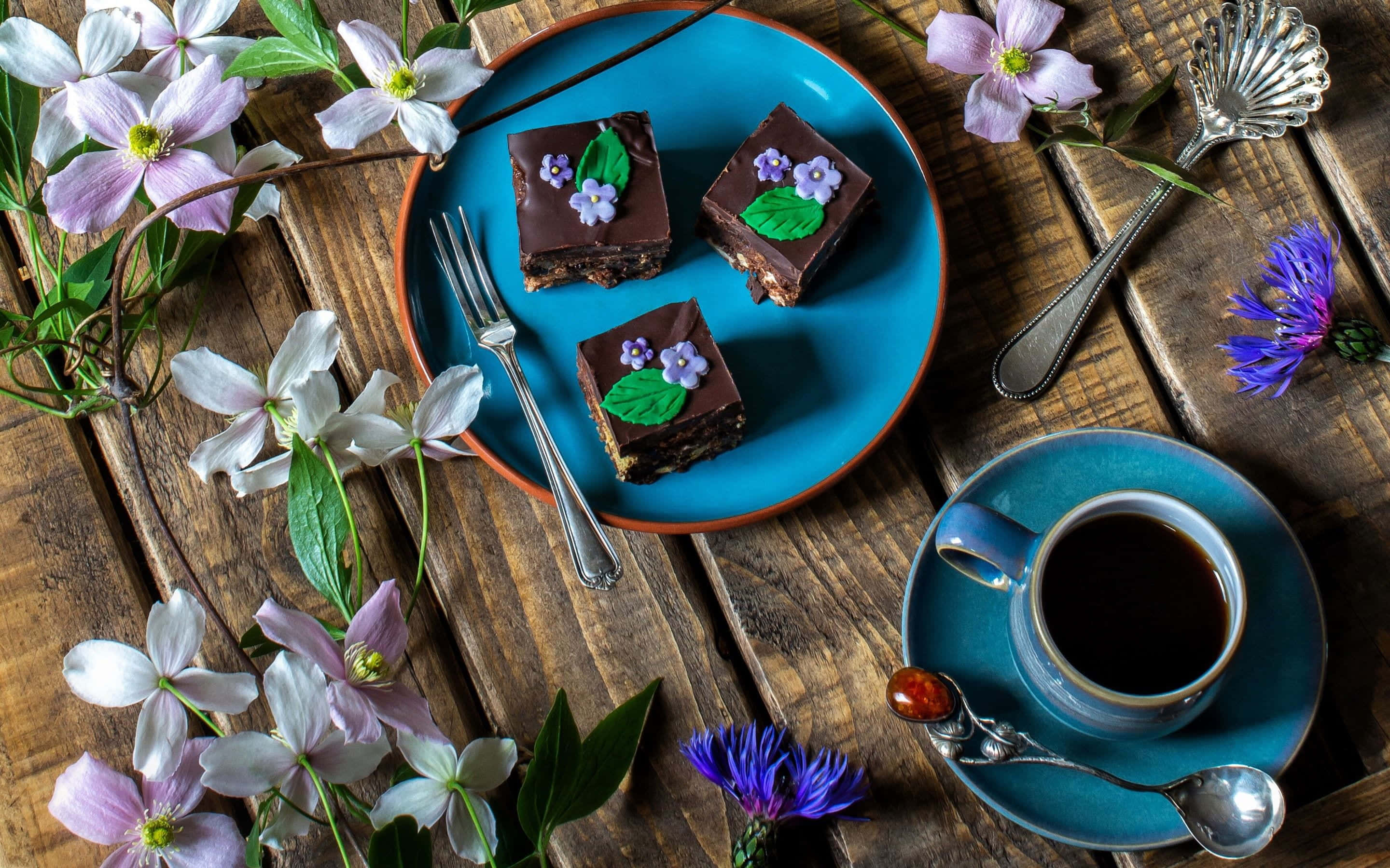 Spring Desserts 桌布 壁紙 — 三塊飾有食用花的朱古力蛋糕方塊放在藍色碟上，旁邊有一杯咖啡，置於鄉村風格的木桌上。