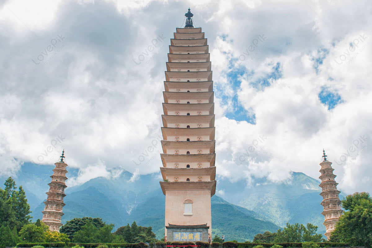 三塔低角度攝影 壁紙 — 三座古老的多層寶塔在戲劇性的多雲天空下聳立，遠處有山巒和下方茂密的綠樹。
