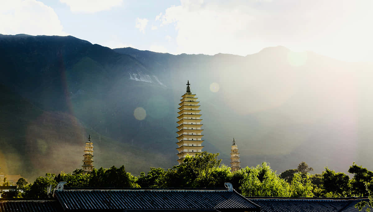 三塔光暈效果 壁紙 — 三座古塔在茂密的綠樹之上聳立，背景是沐浴在金色日出光芒中的霧氣繚繞的山巒。