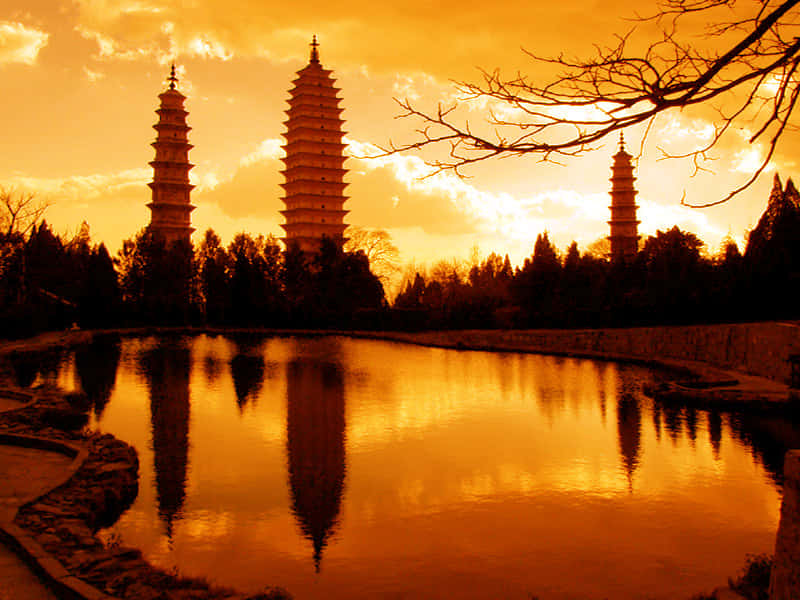 三塔夕陽天 壁紙 — 三座古塔在金色夕陽的映襯下形成剪影，它們的倒影在由光禿樹枝環繞的寧靜池塘中閃爍。