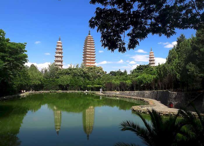 三塔旁的水域 壁紙 — 三座古塔聳立在茂密的綠樹之上，倒映在平靜的池塘中，上方是明亮的藍天。