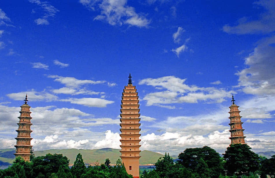 三塔深藍天 壁紙 — 三座古塔雄偉地聳立在鮮豔的藍天下，伴隨著蓬鬆的白雲和遠處的綠色山丘。