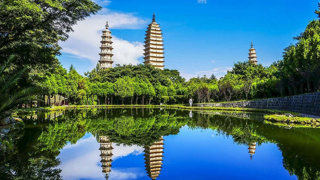 三塔的生動風景 壁紙 — 三座古塔聳立在茂密的綠樹之上，在晴朗的藍天下完美地倒映在平靜的池塘中。
