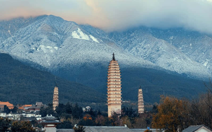 三塔與山景 壁紙 — 三座古塔聳立於城鎮之上，背景是白雪覆蓋的山脈和陰雲密佈的天空。