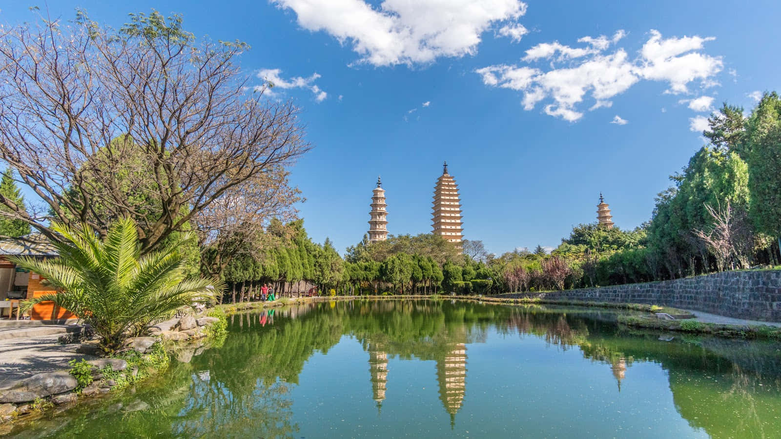三塔與河景 壁紙 — 三座古塔在明亮的藍天下聳立，完美倒映在一個被茂密綠樹環繞的平靜池塘中。