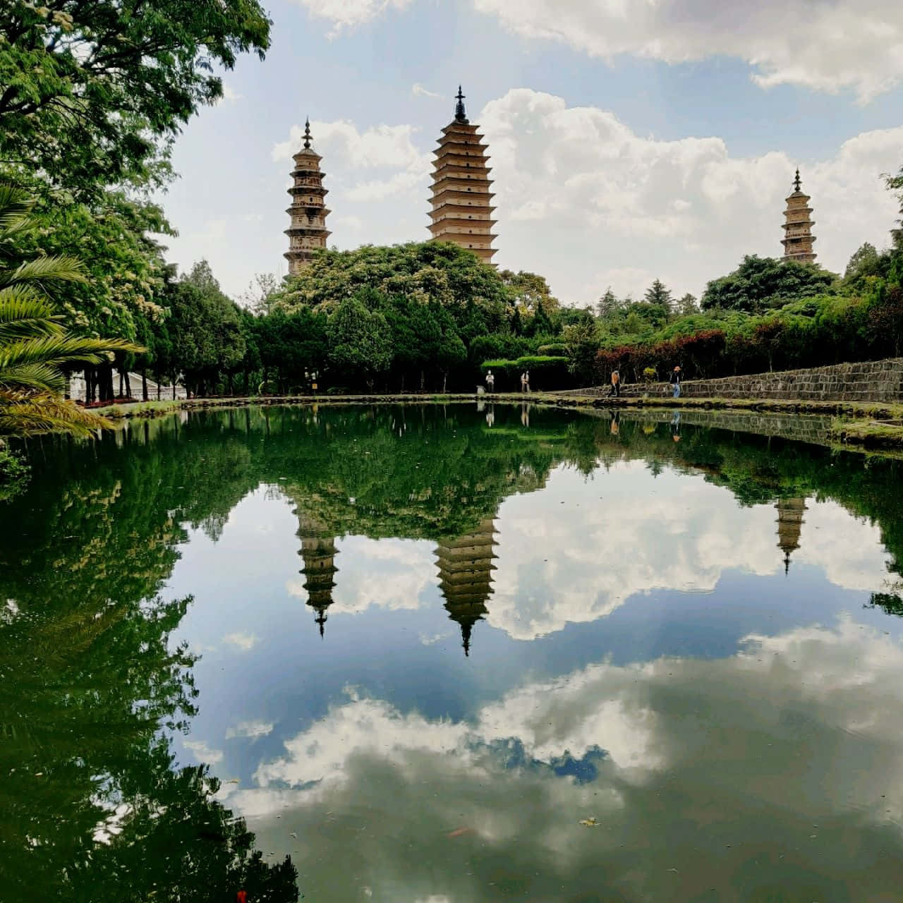 三塔近河景觀 壁紙 — 三座古塔聳立在鬱鬱蔥蔥的綠樹之上，在部分多雲的天空下，完美地倒映在平靜的池塘中。