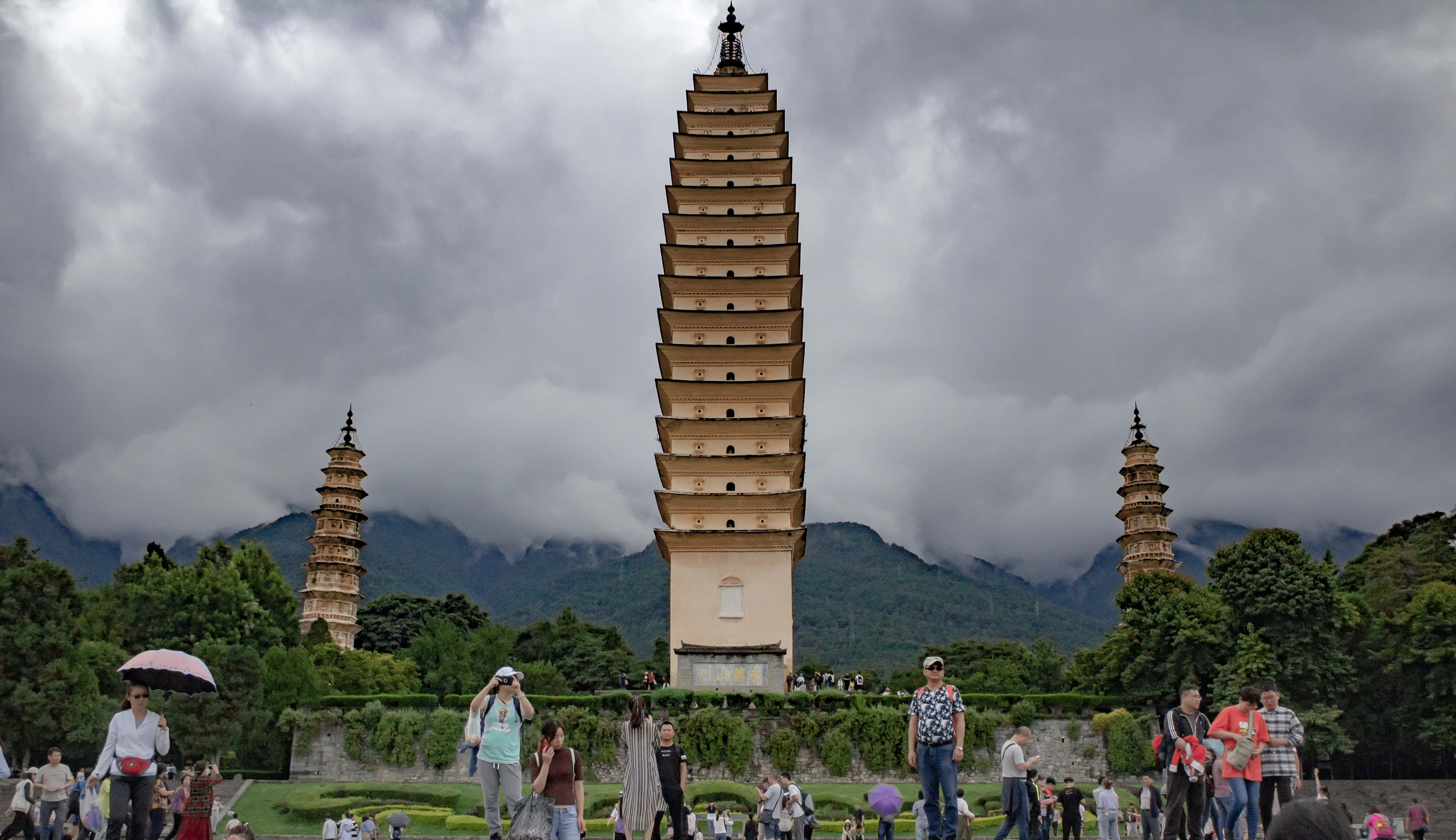 三塔遊人如織 壁紙 — 三座古塔在戲劇性的多雲天空下聳立，背景是霧氣繚繞的山脈，遊客正在探索場地。