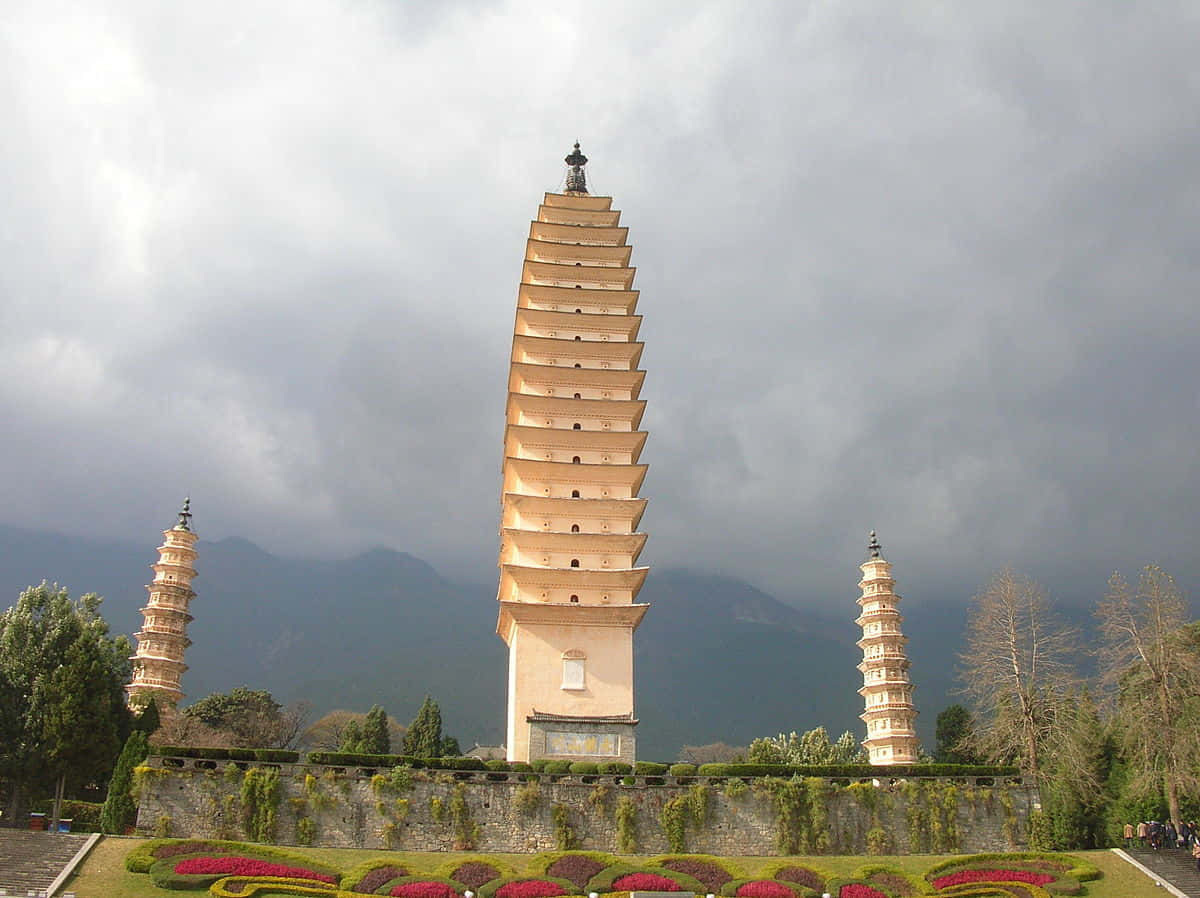 三塔陰雲天 壁紙 — 三層寶塔在戲劇性的多雲天空下聳立，背景是山脈，下方是修剪整齊的花壇。