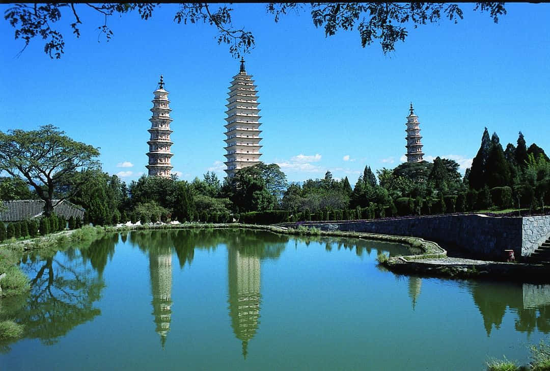 三塔風景 壁紙 — 三座古塔在清澈的藍天下高聳，它們寧靜的倒影完美地映照在一個被茂密綠植環繞的平靜池塘中。
