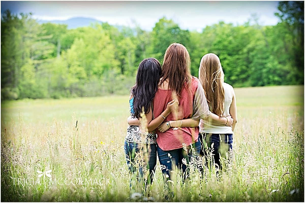 三女生草地合照 壁紙 — 三個女孩在陽光明媚的高草田野中擁抱，背景是樹木繁茂的山丘。