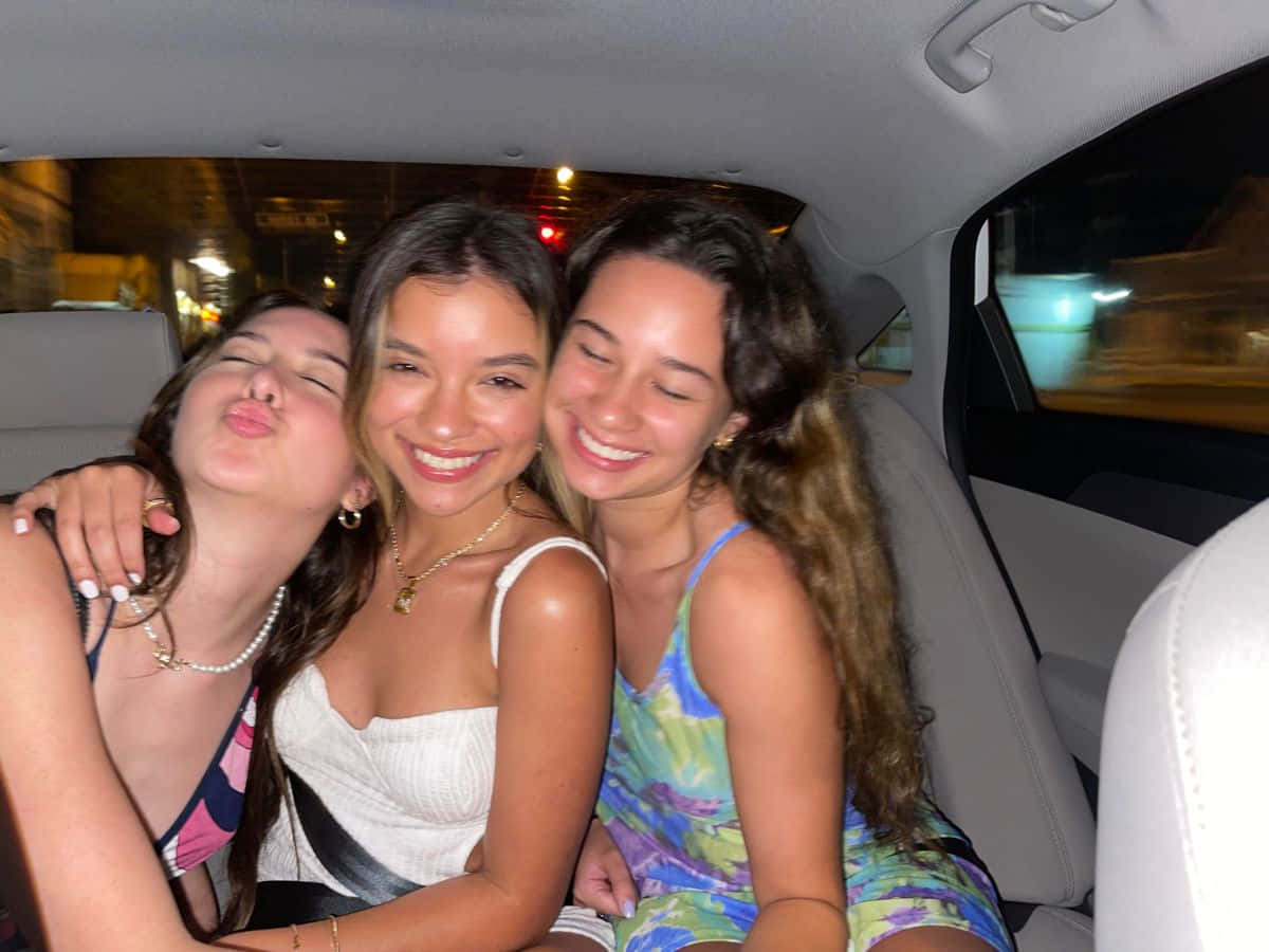 三女生車內合照 壁紙 — 三位微笑的朋友在夜晚的車內頑皮地擺姿勢，模糊的城市燈光從窗外掠過。