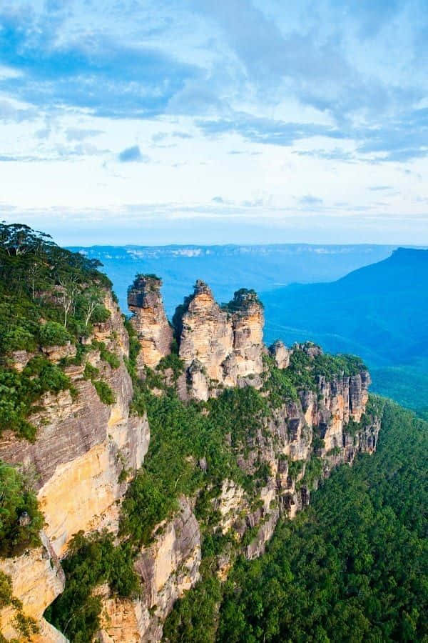 Blue Mountains National Park 桌布 壁紙 — 三座高聳的砂岩柱在明亮的藍天白雲下，從鬱鬱蔥蔥的綠色山谷中戲劇性地升起。