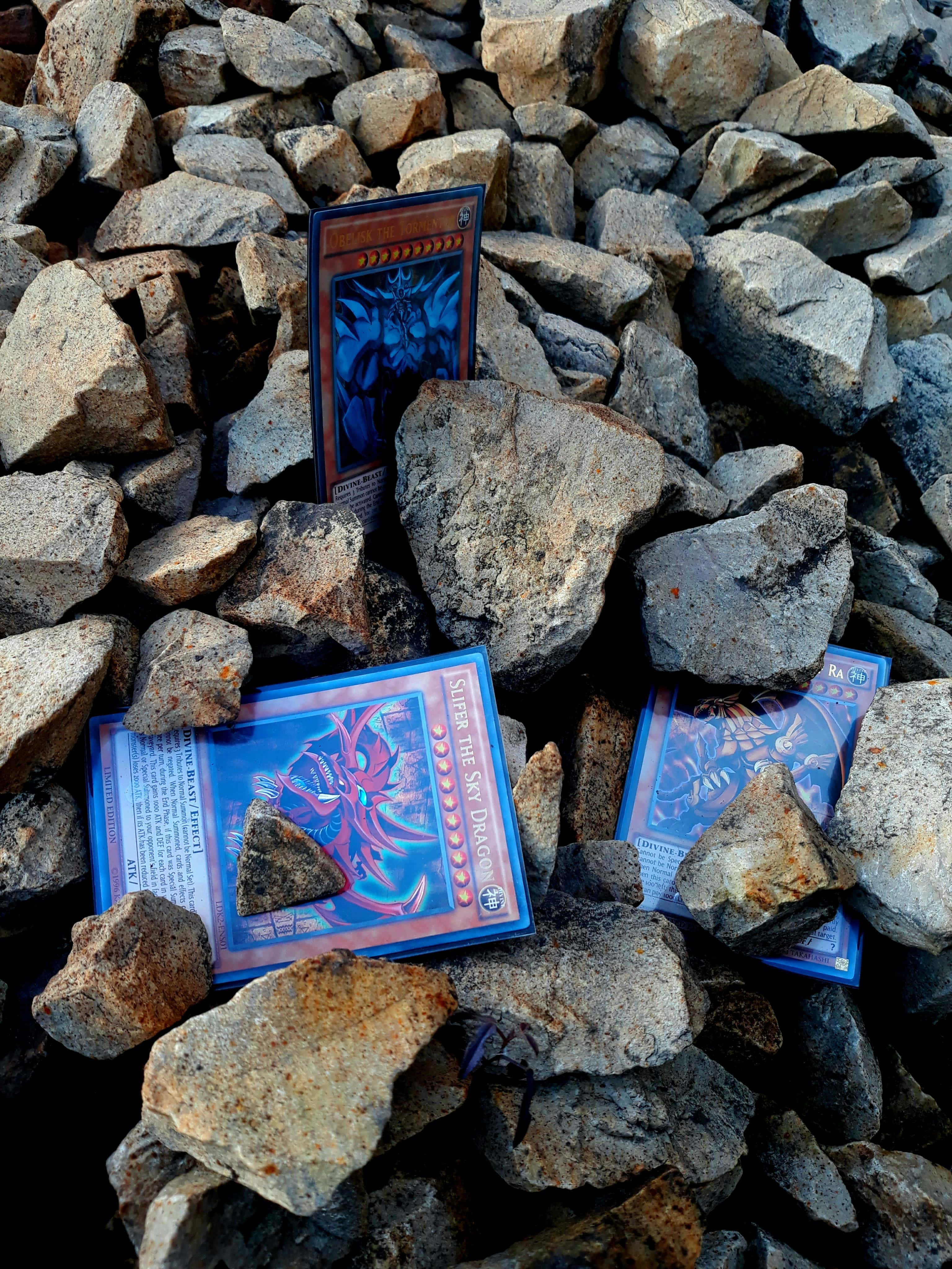 Obelisk The Tormentor 桌布 壁紙 — 三張帶有龍圖案的遊戲王集換式卡牌在明亮的日光下放置在粗糙的灰色岩石之間。
