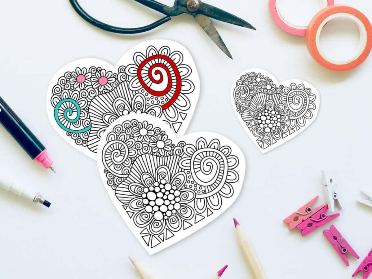 Heart Doodle 桌布 壁紙 — 三張心形填色頁，帶有複雜的漩渦和花朵圖案，周圍環繞著白色表面上的美術用品。