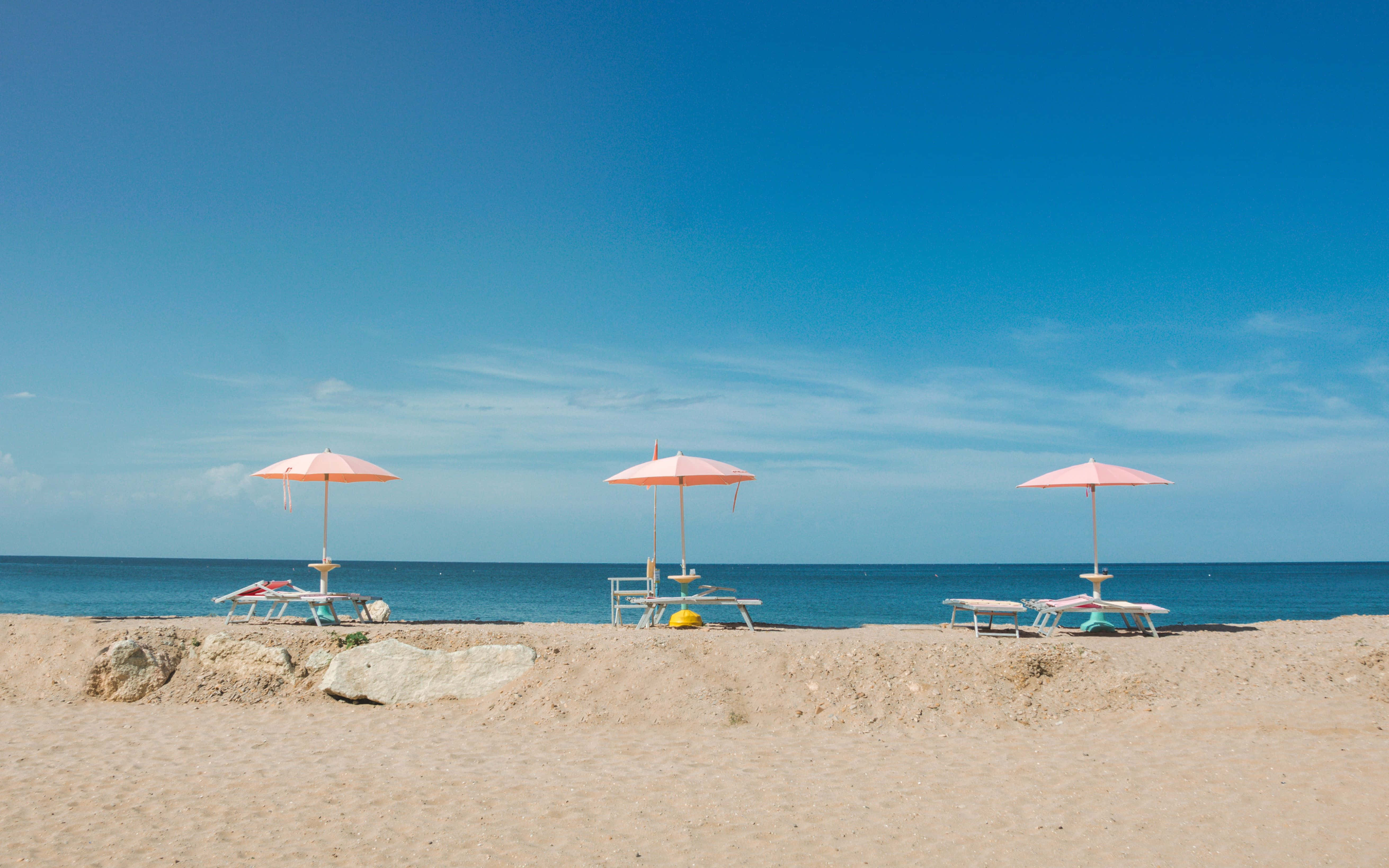 Beach Umbrella 桌布 壁紙 — 三把粉紅色遮陽傘在晴朗的藍天下為沙灘上的空躺椅遮蔭。