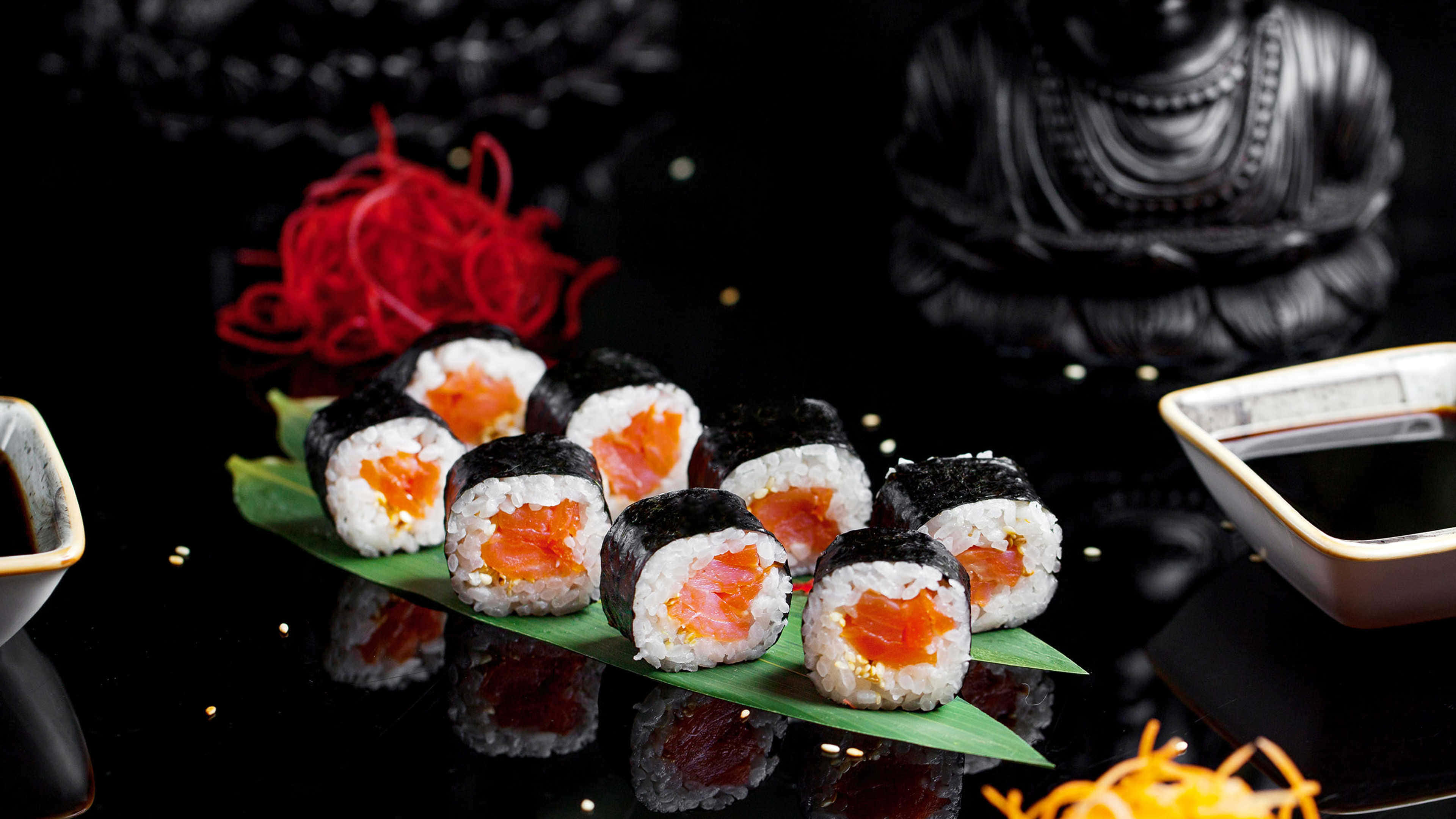Sushi 桌布 壁紙 — 三文魚壽司卷配飯放在綠葉上，置於黑色反光表面，配有紅色裝飾和醬油碗。