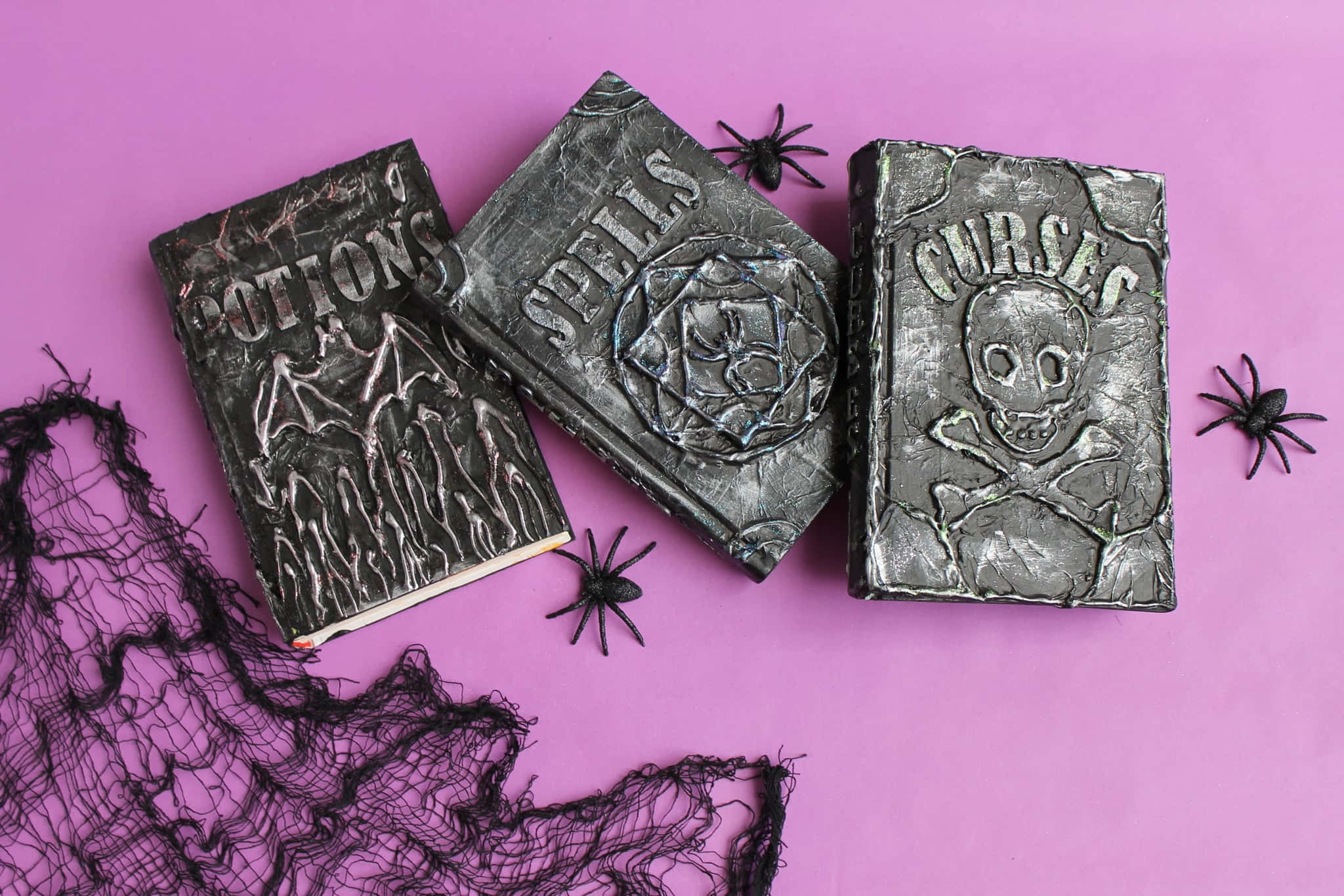 Halloween Books 桌布 壁紙 — 三本帶有紋理的女巫書，標有 Potions Spells Curses，放置在帶有黑色蜘蛛和網狀物的鮮豔粉紅色表面上。