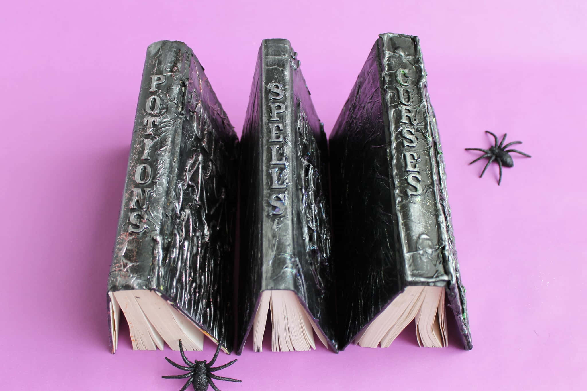 Halloween Books 桌布 壁紙 — 三本殘舊的黑色咒語書，書名分別為《藥水》、《咒語》和《詛咒》，直立於鮮粉紅色表面上，附近有兩隻塑膠蜘蛛。