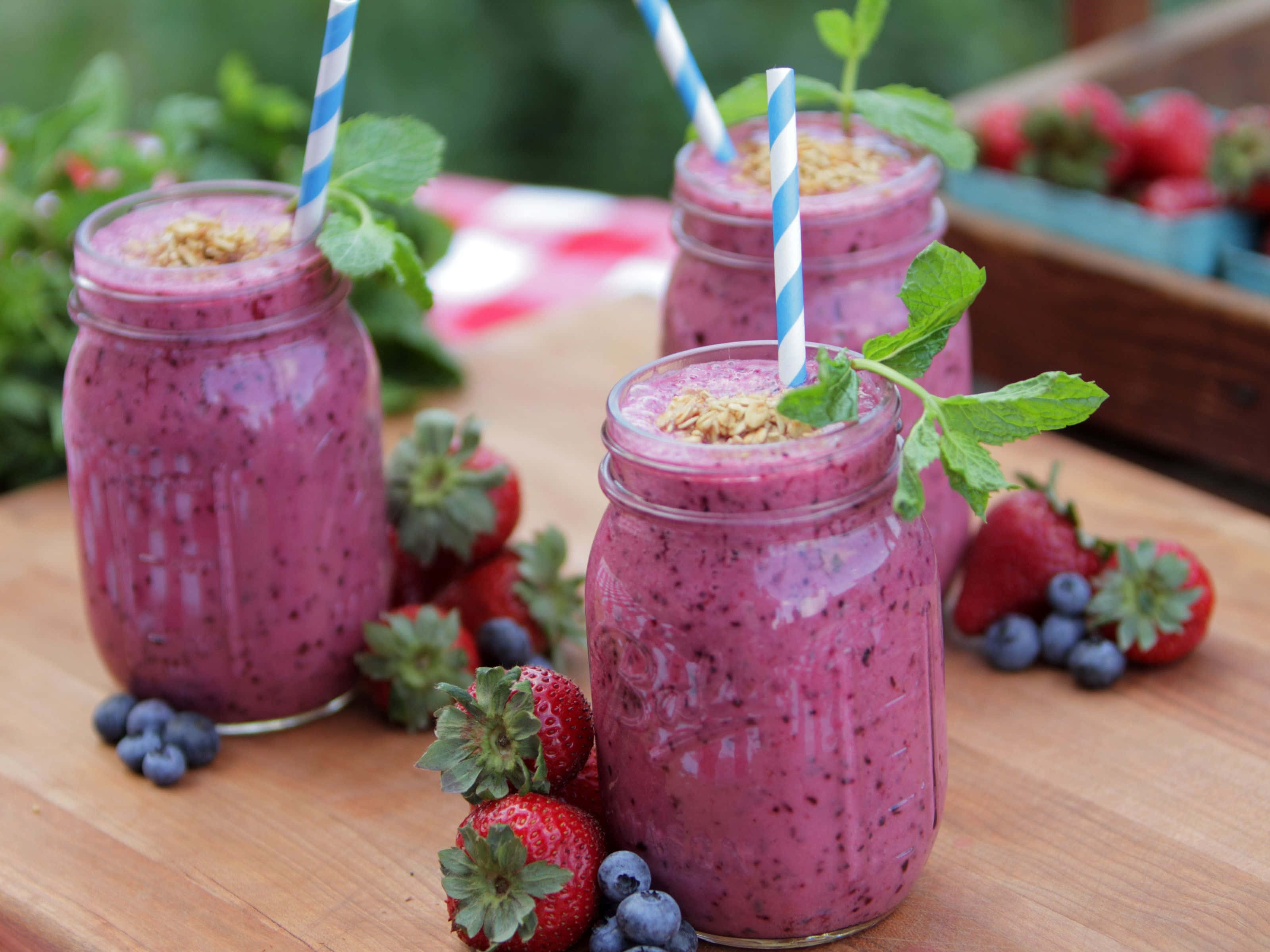 Blueberry Smoothie 桌布 壁紙 — 三杯充滿活力的紫色莓果冰沙裝在梅森罐中，飾以薄荷和格蘭諾拉麥片，放在木板上，周圍是新鮮草莓和藍莓。