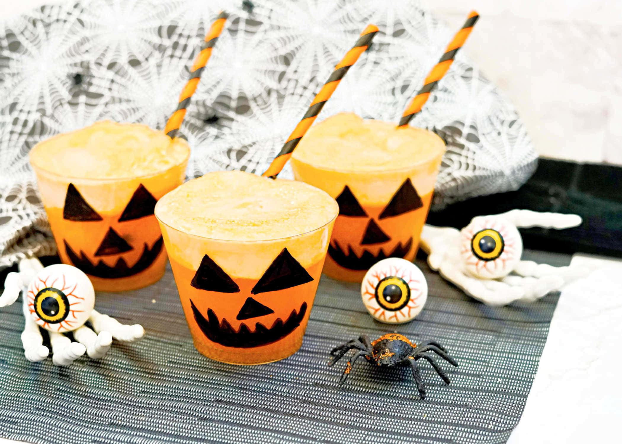 Halloween Drinks 桌布 壁紙 — 三杯橙色南瓜臉飲品，附有條紋吸管，放在灰色墊子上，周圍有假眼球和一隻蜘蛛。