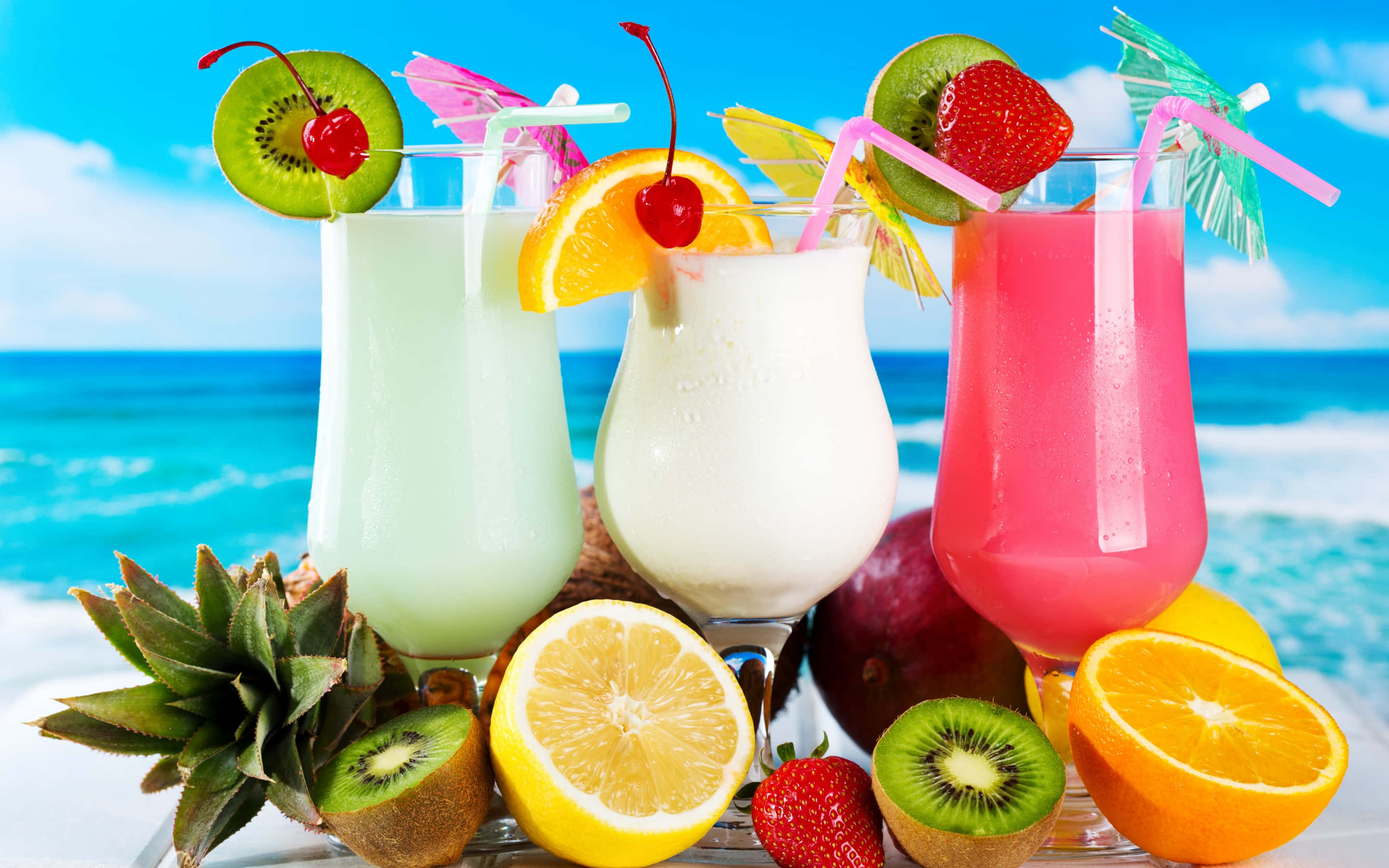 Food And Beverage 桌布 壁紙 — 三杯熱帶水果飲品，飾以奇異果、櫻桃和橙，放在沙灘桌上，旁邊有切片柑橘和菠蘿，背景是明亮的藍色海洋。