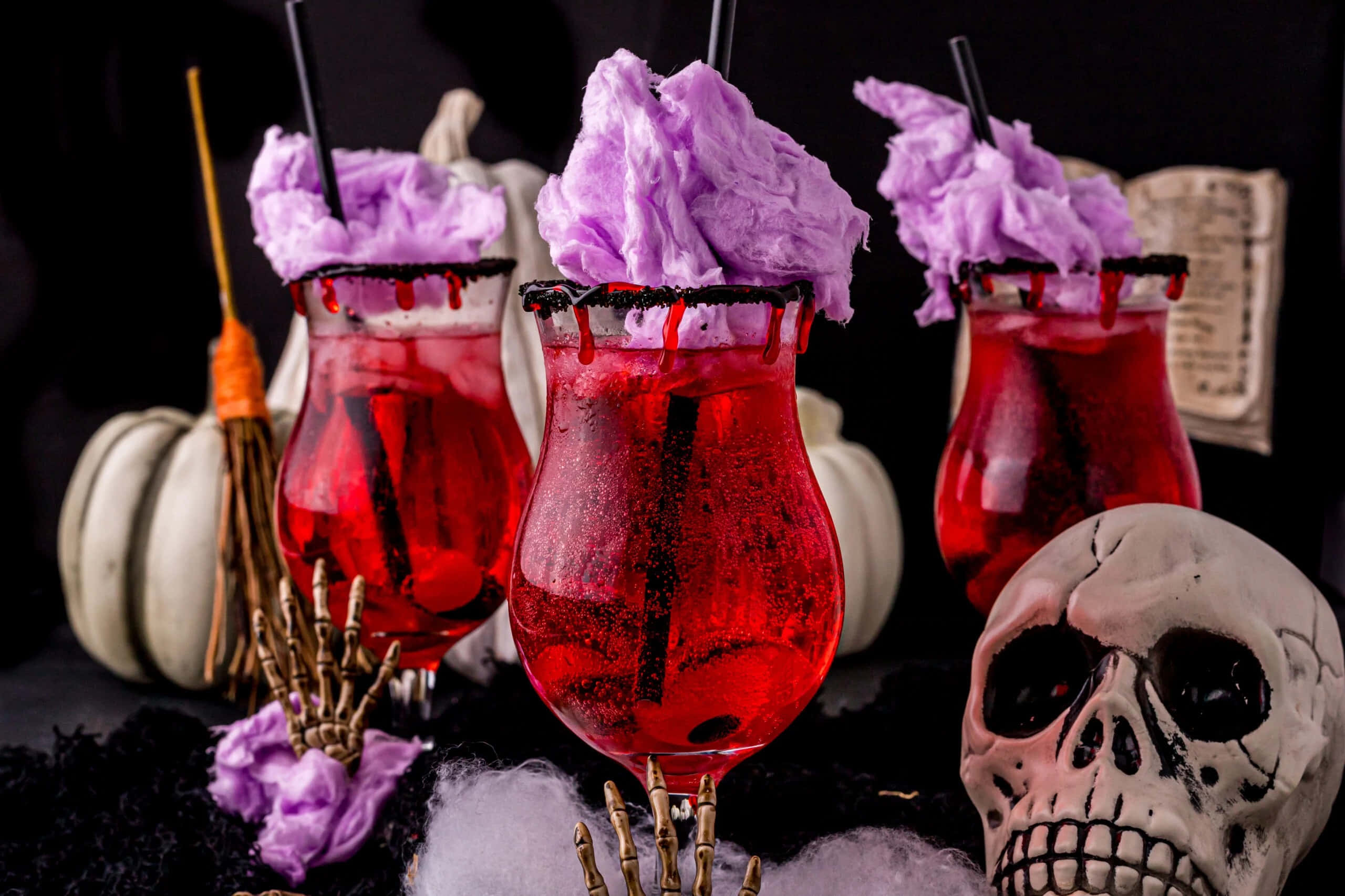 Halloween Drinks 桌布 壁紙 — 三杯紅色萬聖節雞尾酒，上面有紫色棉花糖和黑色糖邊，旁邊放著一個骷髏頭和南瓜。