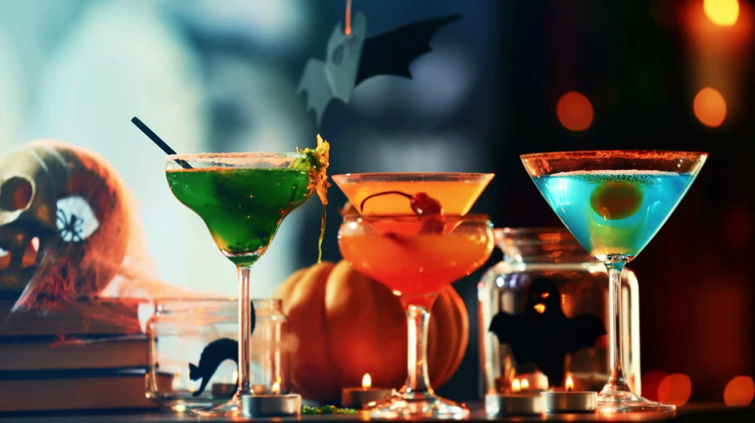 Spooky Cocktails 桌布 壁紙 — 三杯色彩繽紛的萬聖節雞尾酒盛載於馬提尼杯中，放在桌上，旁邊有南瓜和鬼怪裝飾，在溫暖的燭光下。