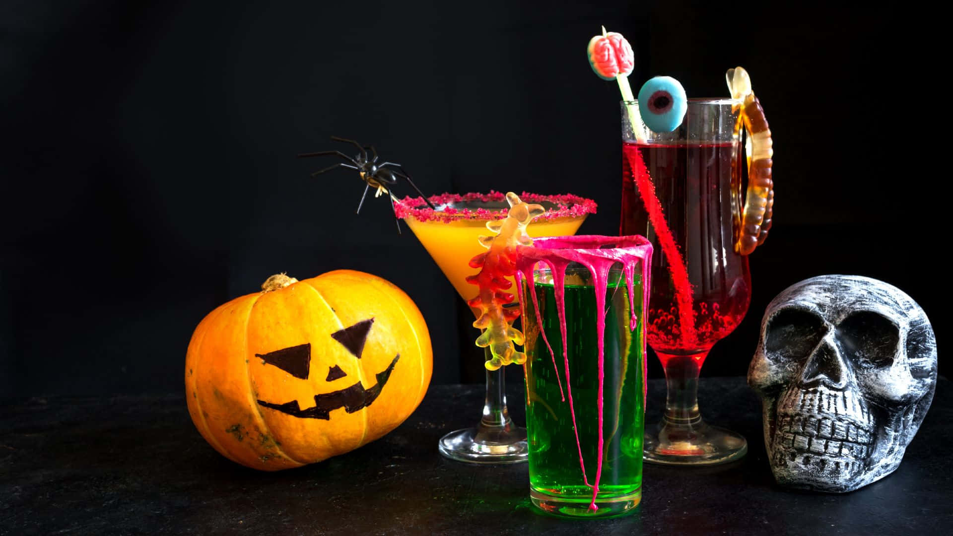 Spooky Cocktails 桌布 壁紙 — 三杯詭異的萬聖節雞尾酒，配上糖果裝飾，放在一個雕刻南瓜和骷髏旁邊，背景為黑色。