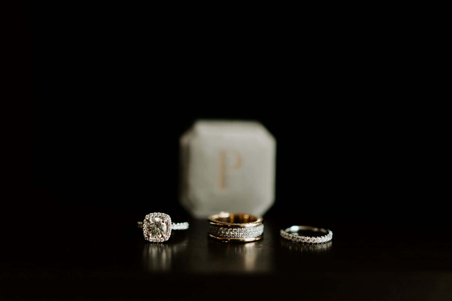 Wedding Rings 桌布 壁紙 — 三枚優雅的鑽石戒指在深色表面上閃耀，背景有一個模糊的白色戒指盒，上面標有「P」。