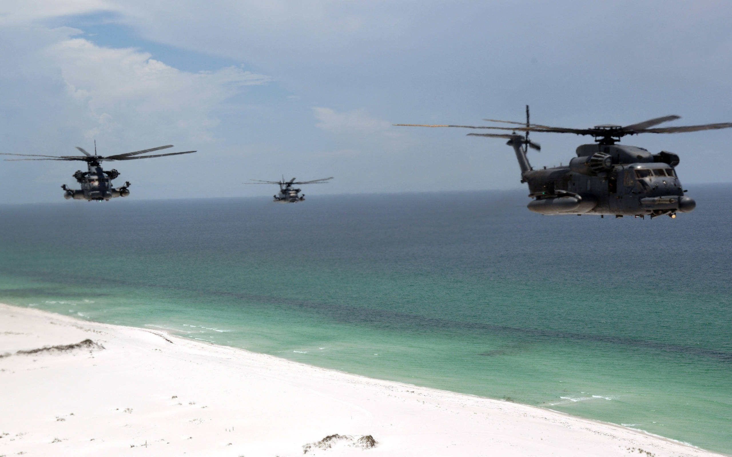 三架Sikorsky MH-53軍用直升機 壁紙 — 三架軍用直升機低飛過一片純淨的白色沙灘，與碧綠色的海洋相接，天空部分多雲。