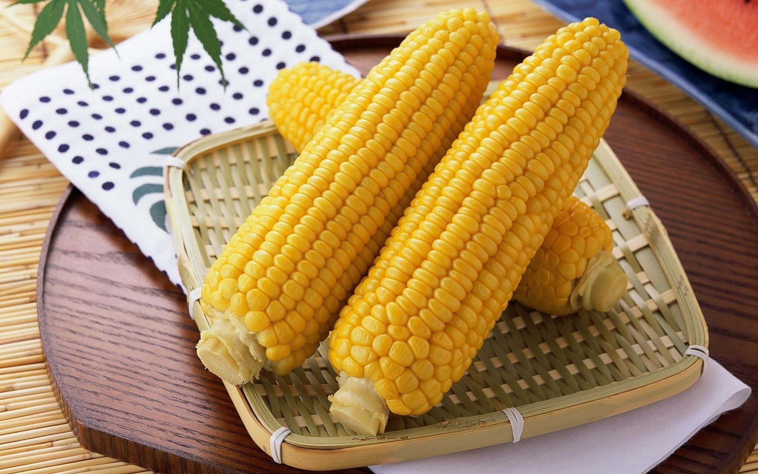 Yellow Corn 桌布 壁紙 — 三條鮮黃色的粟米放在木托盤上的編織竹籃中，旁邊有一片西瓜和葉狀裝飾。