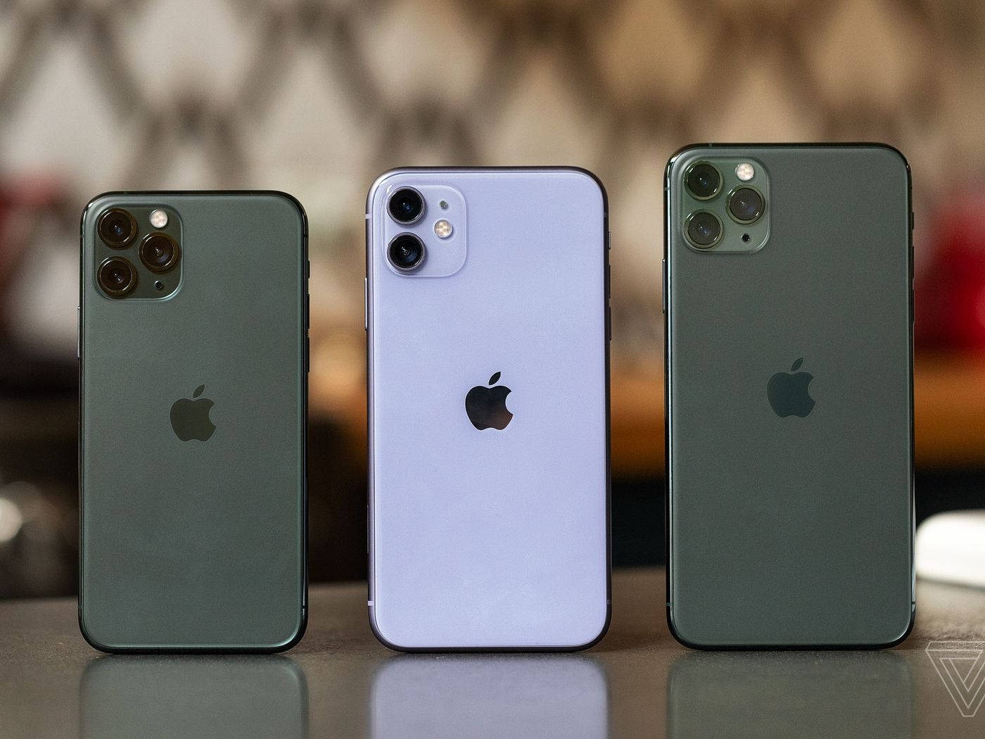 三款 iPhone 11 系列 壁紙 — 三款綠色和紫色的Apple iPhone並排放在反光表面上，背景是模糊的圖案。