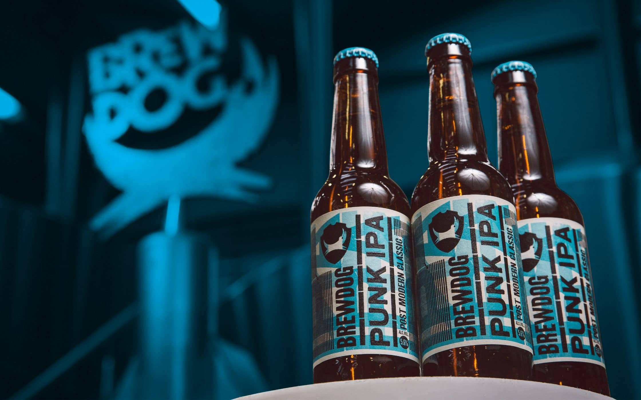 三瓶 BrewDog Punk Ipa 壁紙 — 三支 BrewDog Punk IPA 啤酒瓶在發光的藍色 BrewDog 標誌背景前清晰對焦。