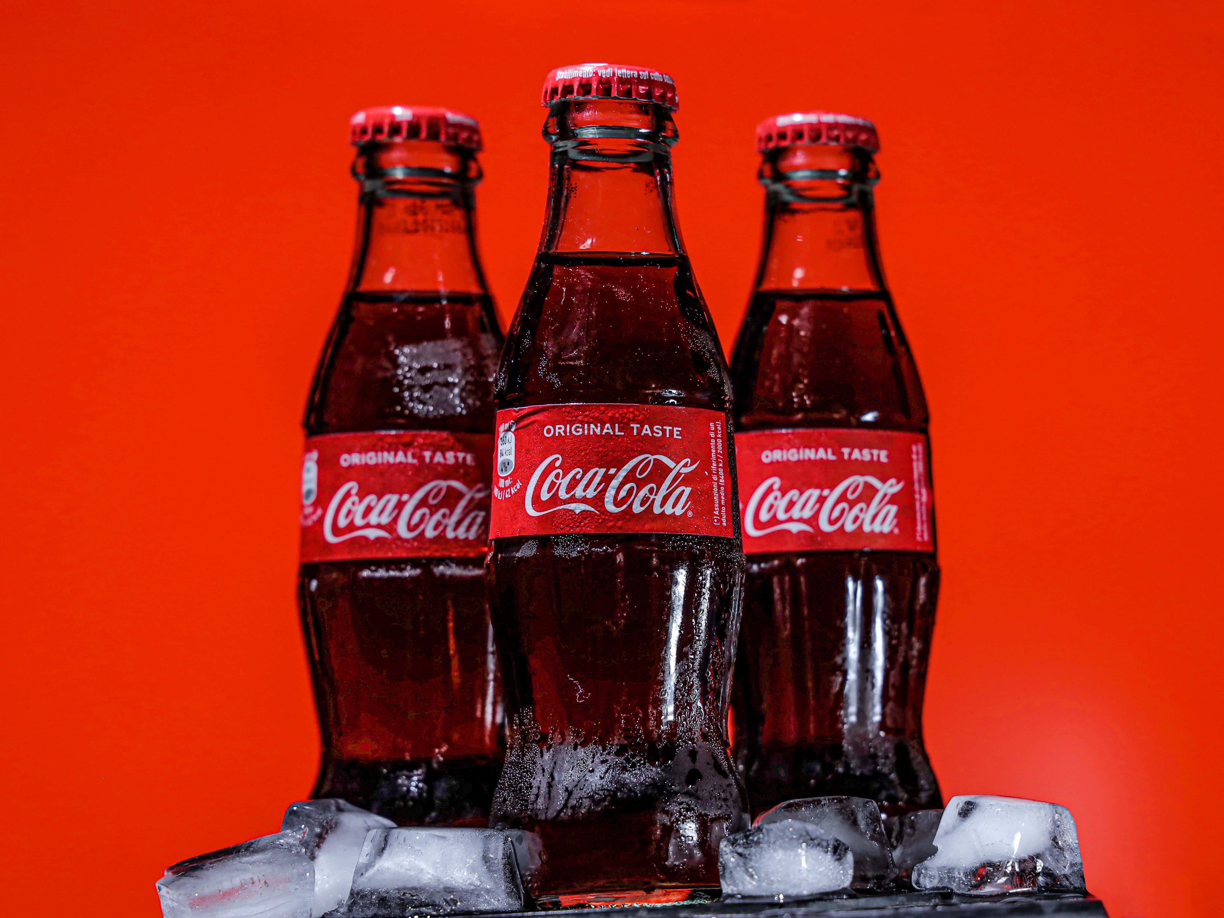 coca cola 桌布 壁紙 — 三瓶冰凍的可口可樂玻璃瓶，帶有冷凝水，放在冰上，背景為鮮紅色。