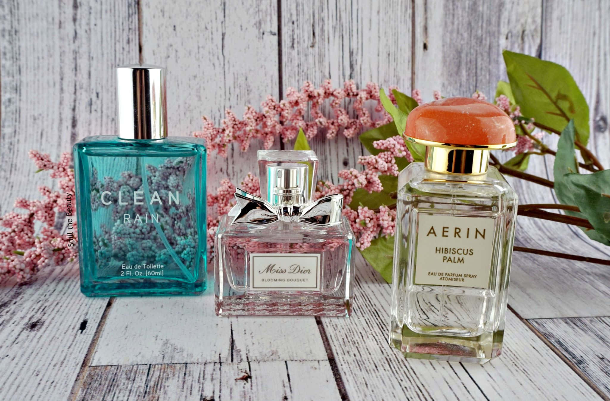 Spring Perfume 桌布 壁紙 — 三瓶標有 Clean Rain、Miss Dior 和 Aerin Hibiscus Palm 的香水瓶放在風化木上，旁邊有粉紅色的花朵。