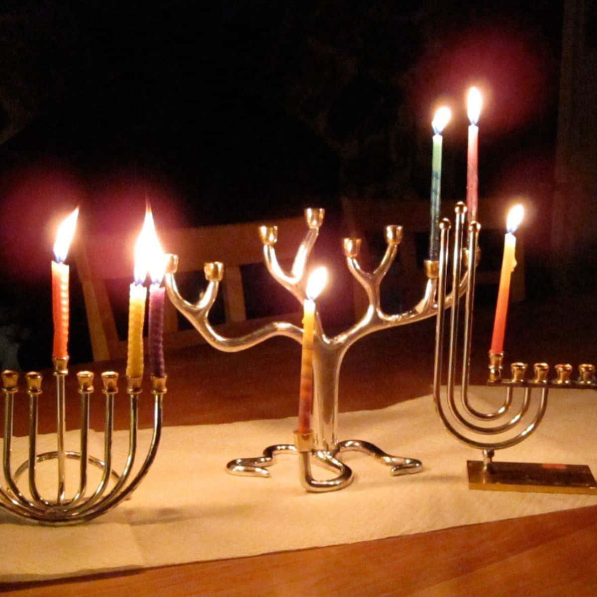 Hanukkah 桌布 壁紙 — 三盞點亮的燭台（Menorah）在黑暗的夜晚背景下，桌上彩色蠟燭溫暖地閃耀。