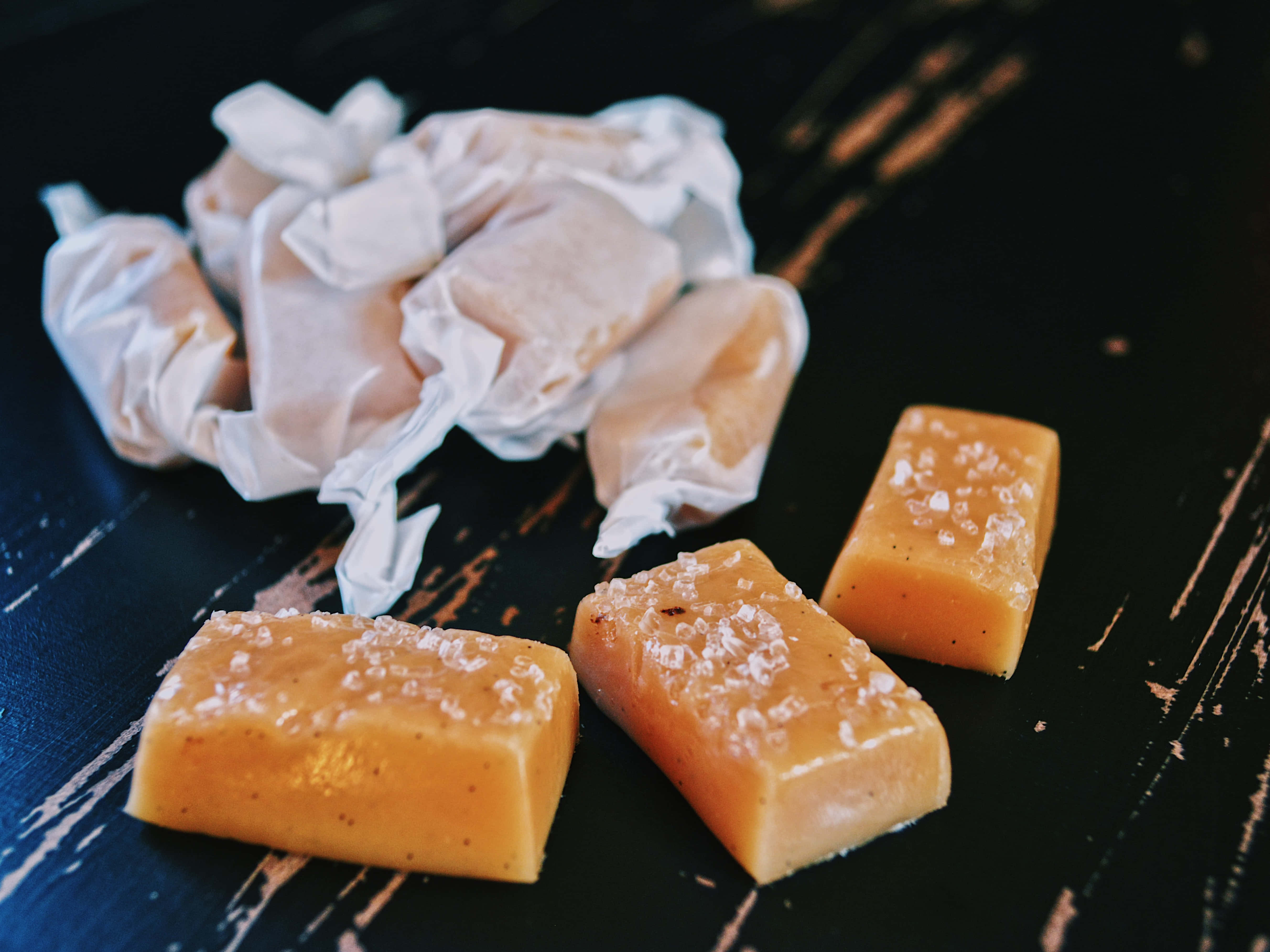 Caramel 桌布 壁紙 — 三粒海鹽焦糖糖果，帶有粗糖，放在深色鄉村風木面上，旁邊有皺巴巴的包裝紙。