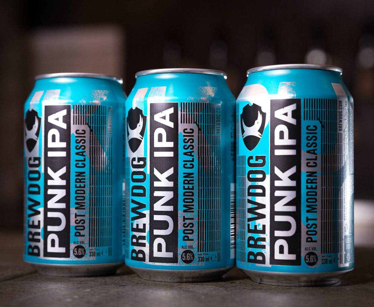 三罐 BrewDog Punk IPA 啤酒罐於深色背景 壁紙 — 三罐亮藍色嘅BrewDog Punk IPA啤酒罐排列喺深色木質表面上，景深較淺。