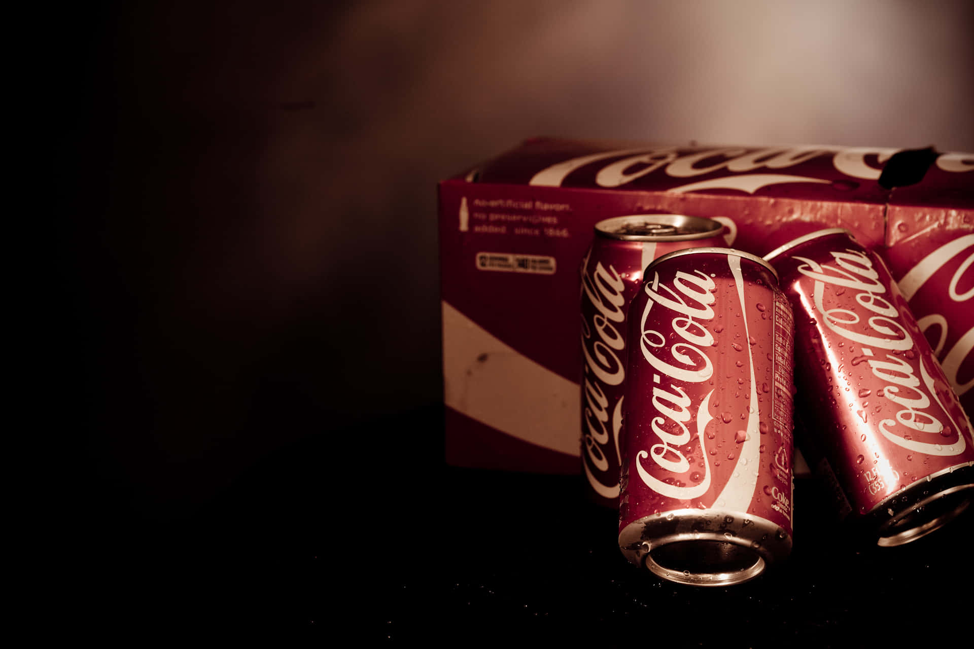 coca cola 桌布 壁紙 — 三罐冰凍的可口可樂罐，帶有水珠，放在紅色紙板架旁，背景陰暗有氣氛。