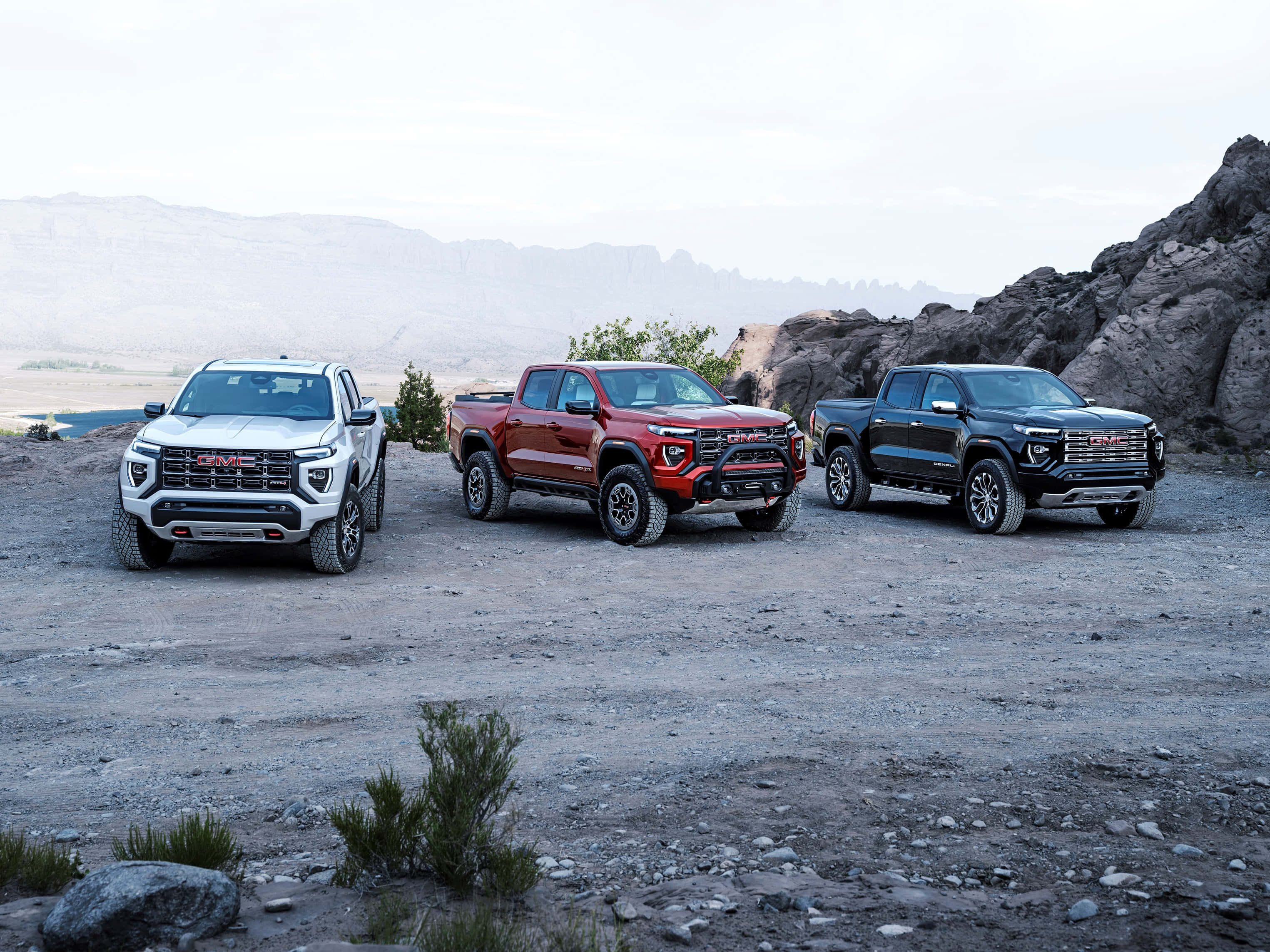 Gmc Canyon 桌布 壁紙 — 三輛堅固的GMC皮卡車，分別為銀色、紅色和黑色，並排停放在岩石沙漠地形上，背景是遠山和陰天。