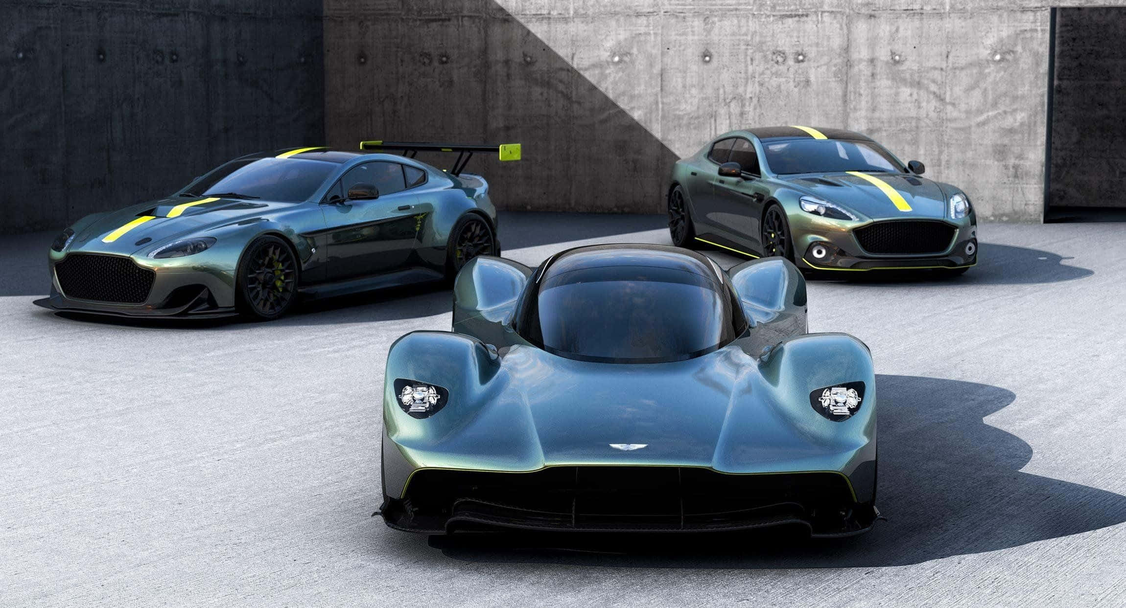 Aston Martin Valkyrie 桌布 壁紙 — 三輛時尚的Aston Martin超級跑車，金屬綠色配黃色條紋，停在灰色牆壁前的混凝土上。