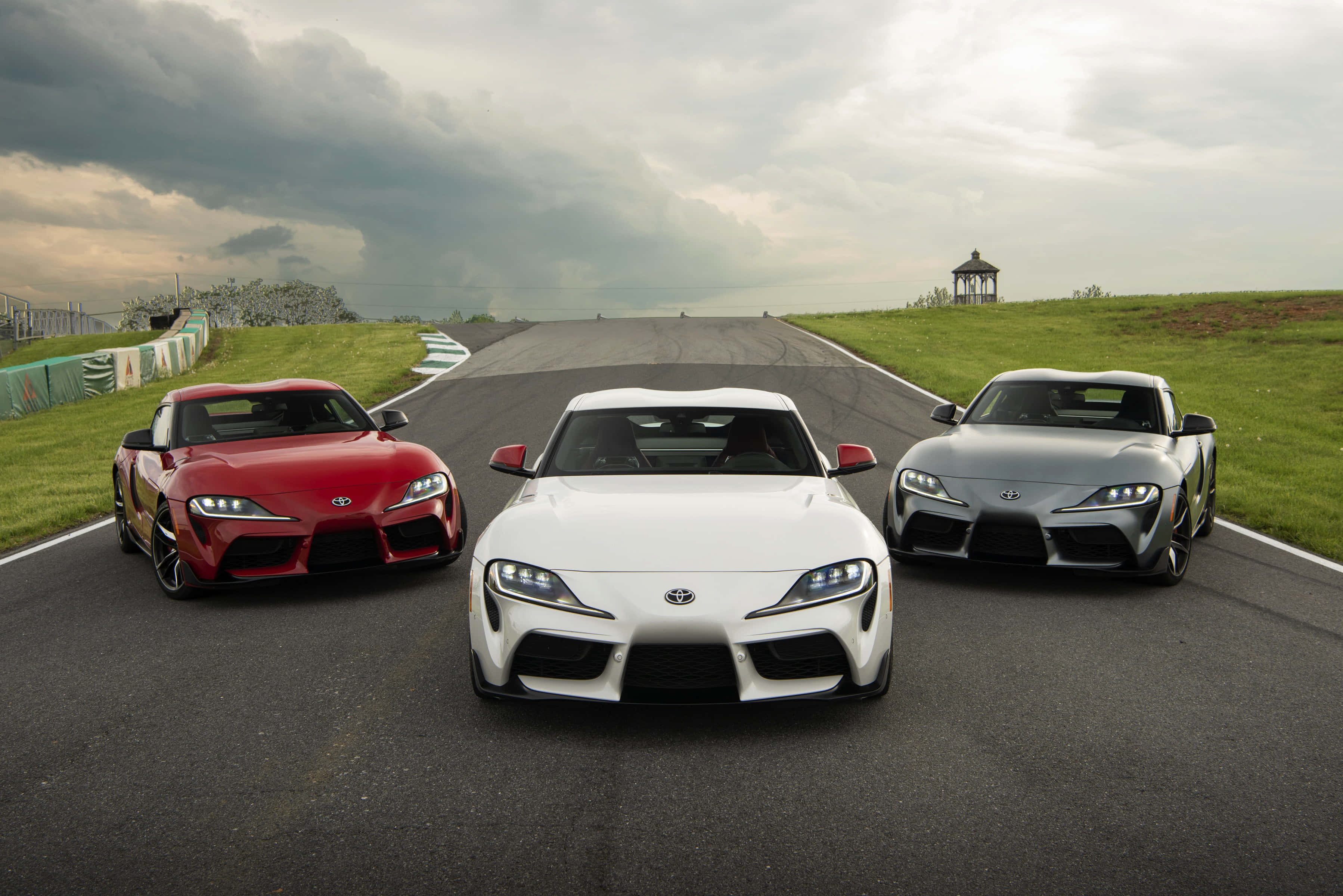 Supra 桌布 壁紙 — 三輛流線型豐田Supra跑車（紅、白、銀）在暴風雨天空下的賽道上蓄勢待發。