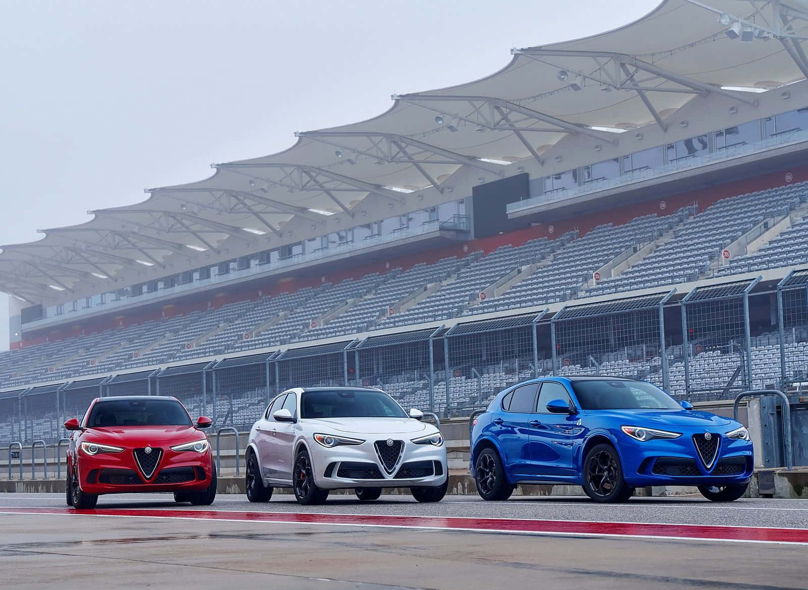 Alfa Romeo Stelvio 桌布 壁紙 — 三輛紅色、白色和藍色的愛快羅密歐SUV在陰天下一字排開在賽道上，後面是空無一人的看台。